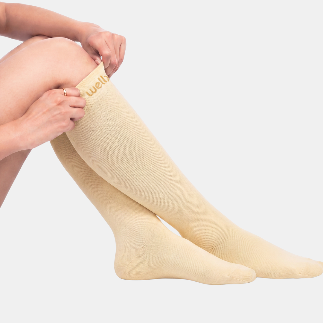 Candlelight Yellow Compression Socks For women, A person wearing light beige over-the-knee ribbed socks, with one hand adjusting the top cuff. The brand name 'well.