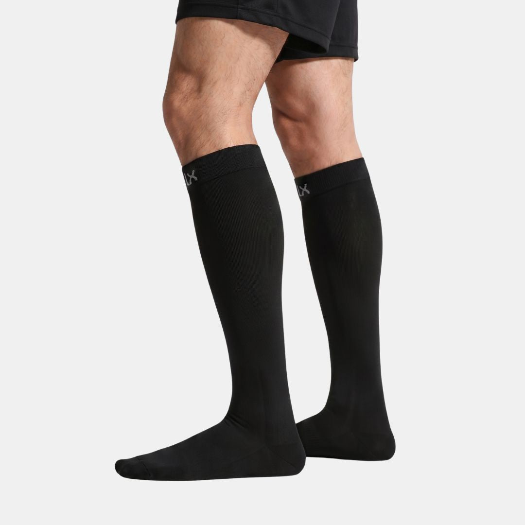 Black Compression Socks for Men, A person's legs wearing black low-cut athletic socks with a small white logo on the Cuff, paired with black athletic shorts, against a light gray studio background.