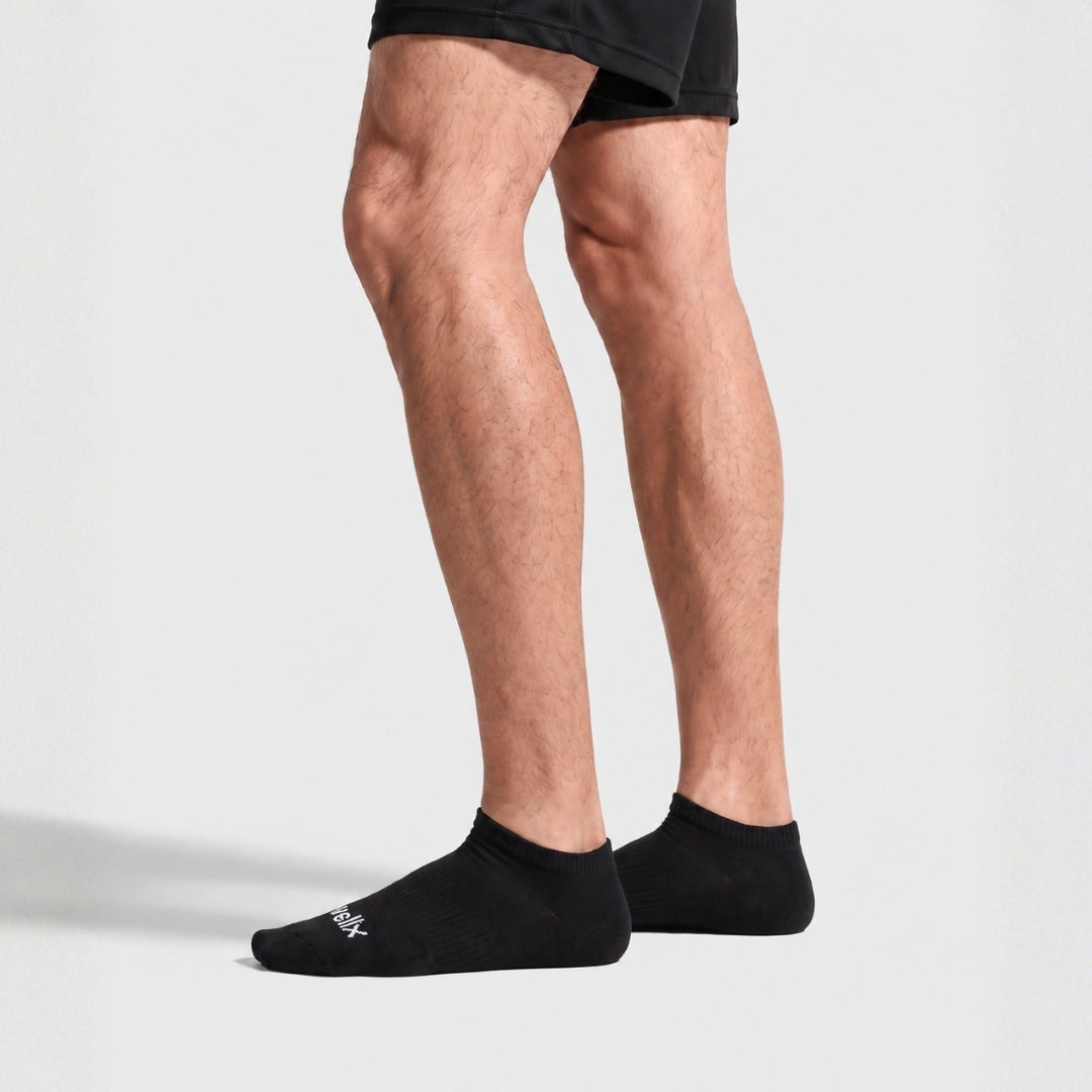 Black Street Ankle Socks For Men. A person's legs wearing black low-cut athletic socks with a small white logo on the toe, paired with black athletic shorts, against a light gray studio background.