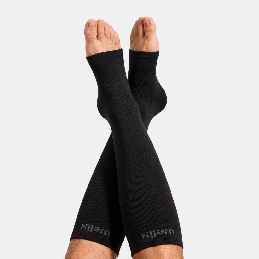 Black Toeless Compression Socks For Men Black compression sleeves worn on crossed legs, with the Wellx brand name visible near the lower cuffs, against a light gray background.