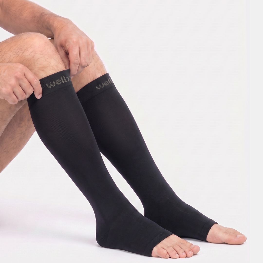 Black Toeless Compression Socks For Men A person seated against a light gray studio background wearing black knee-high compression sleeves. The sleeves extend from the upper leg down to the foot, and the brand name 'wellx' is visible in gray on the cuffs.