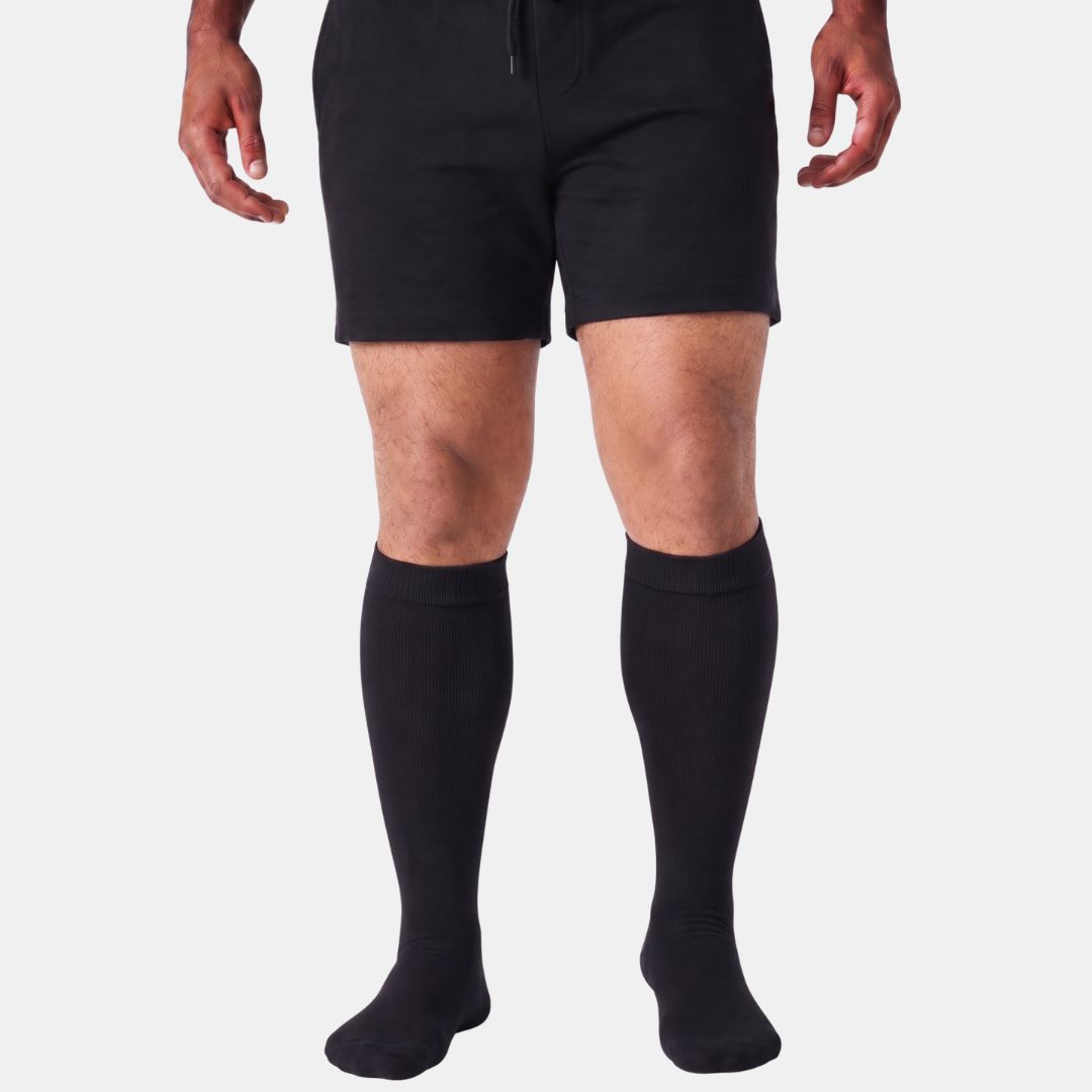 Black Wide Calf Compression Socks For Men. A person from the waist down wearing black athletic shorts and matching black knee-high socks against a light gray studio background.