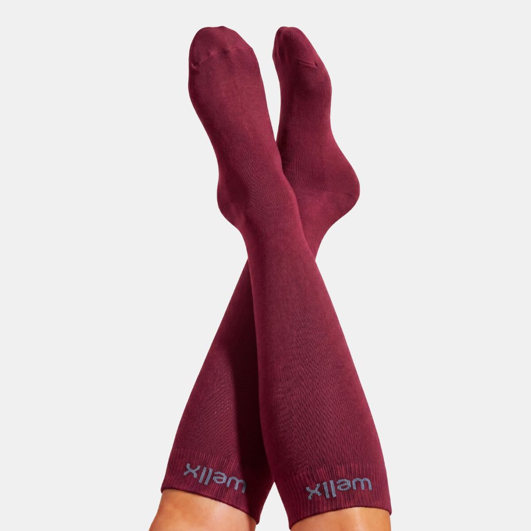 Burgundy Compression Socks For Men, A pair of maroon crew socks worn on crossed legs, with the brand name 'wellx' visible in light gray at the ribbed cuffs, against a solid light gray background.