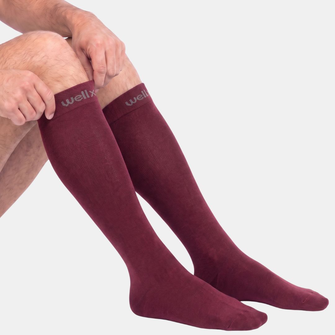 Burgundy Compression Socks For Men, A close-up of a person's legs wearing burgundy over-the-knee socks with the brand name 'wellx' visible on the top cuff, set against a plain light gray background.