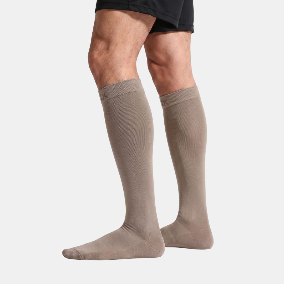 Gravel Compression Socks For Men. A person's legs wearing a pair of knee-high taupe socks, shown from the mid-thigh down against a light gray studio background.