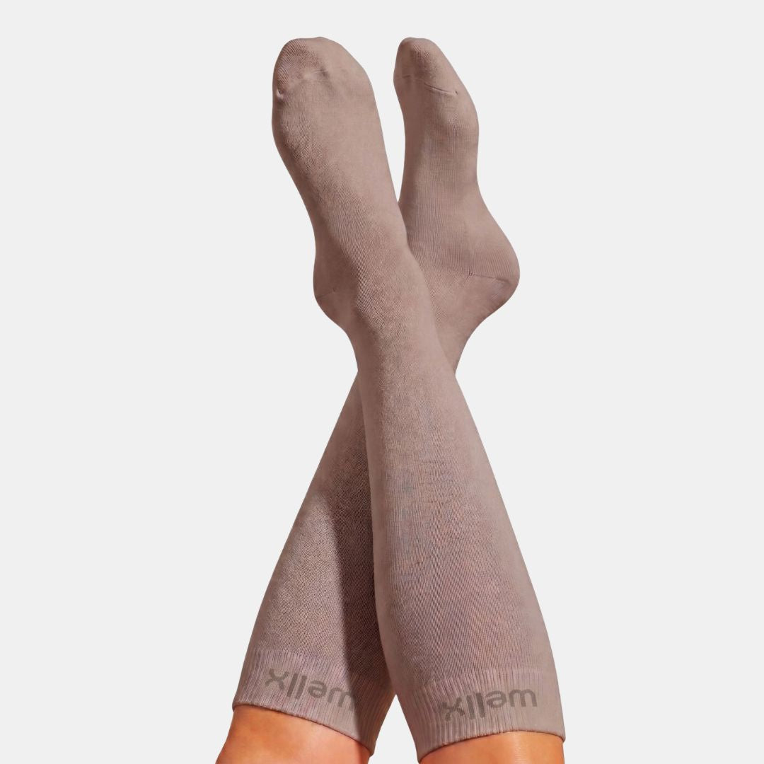 Gravel Compression Socks For Men. A pair of ribbed knee-high socks in a muted taupe-brown color, worn on legs with the feet raised against a light gray background. The brand name 'wellx' is visible on the ribbed cuffs.