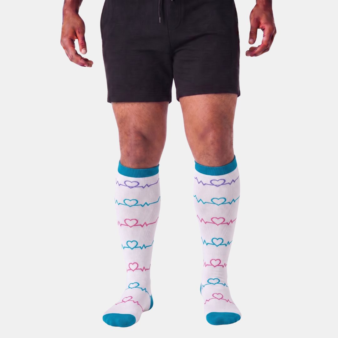 Heartbeat White Wide Calf Compression Socks For Men. A person from waist to feet wearing knee-high socks with a white base, blue cuffs and toes, and pink and blue heartbeat line patterns, paired with black shorts against a light gray background.