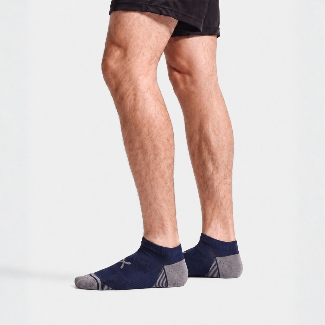 Navy Power Stride Ankle Socks Form Men. A person's legs wearing navy blue and gray low-cut athletic socks, paired with black athletic shorts, against a light gray studio background.