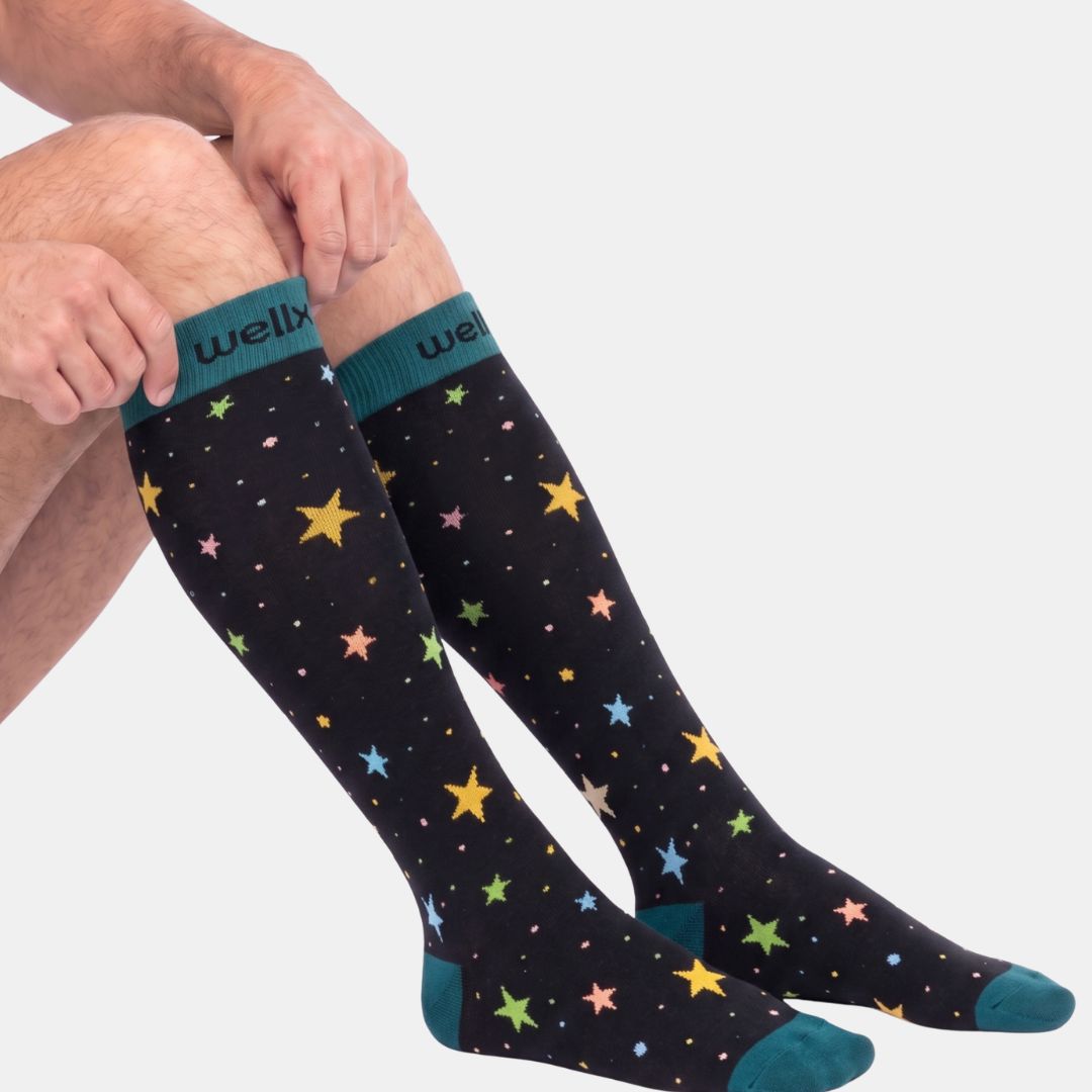 Stars Black/Green Compression Socks for Men. A person wearing black knee-high socks with a colorful star pattern and teal cuffs, heels, and toes. The brand name 'wellx' is visible on the cuff.