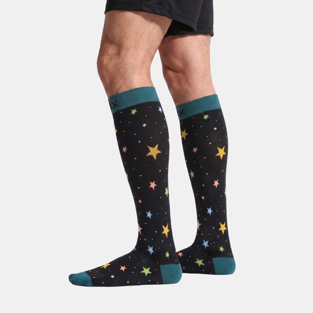 Stars Black/Green Compression Socks for Men. A person's legs wearing a pair of black knee-high socks with a colorful star pattern and teal cuffs, heels, and toes, shown against a light gray background.