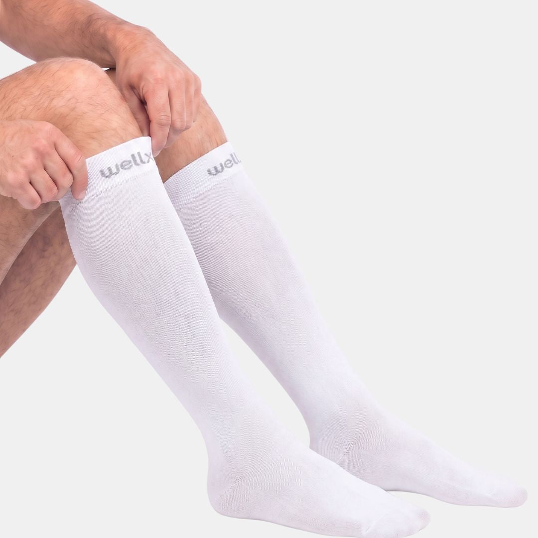 Compression Socks For Men, White knee-high compression socks worn on a person's legs, with hands adjusting one sock against a light gray studio background. The brand name 'well.