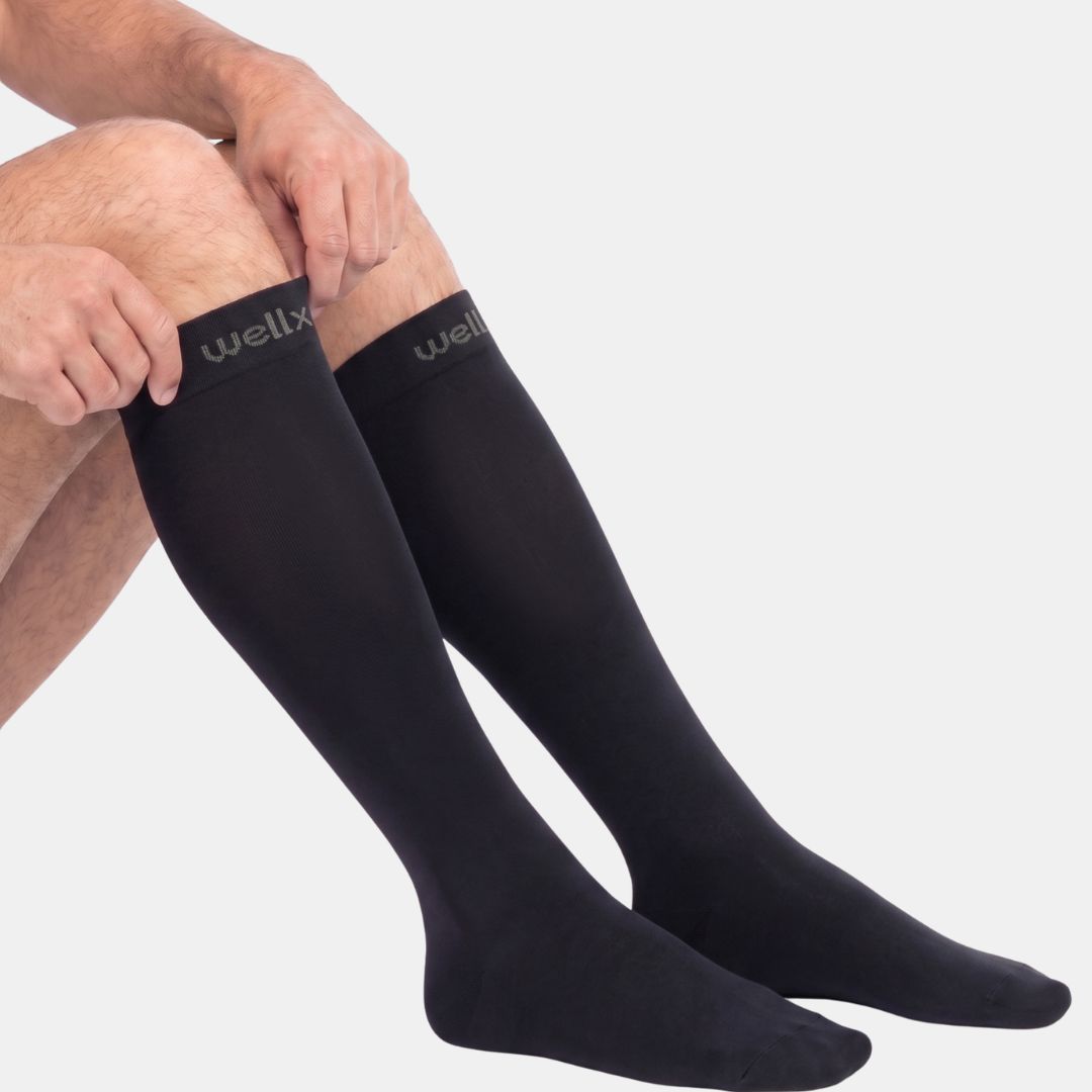 Black Compression Socks for Men A person's legs wearing black low-cut athletic socks with a small white logo on the Cuff, paired with black athletic shorts, against a light gray studio background.