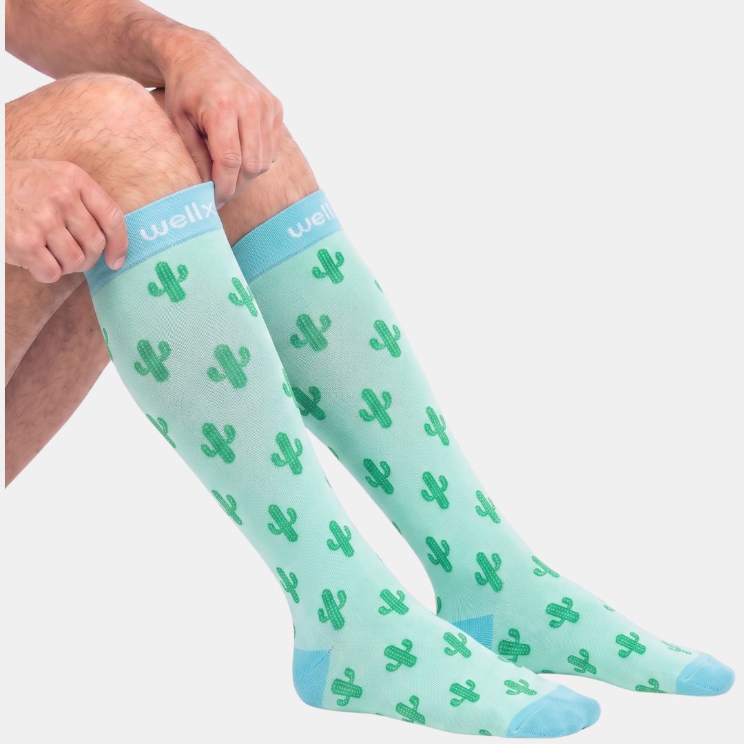 Cactus Green Compression Socks For Men, A person's legs wearing knee-high socks with a green cactus pattern on a mint green background, with a light blue cuff and toes. The brand 'well.