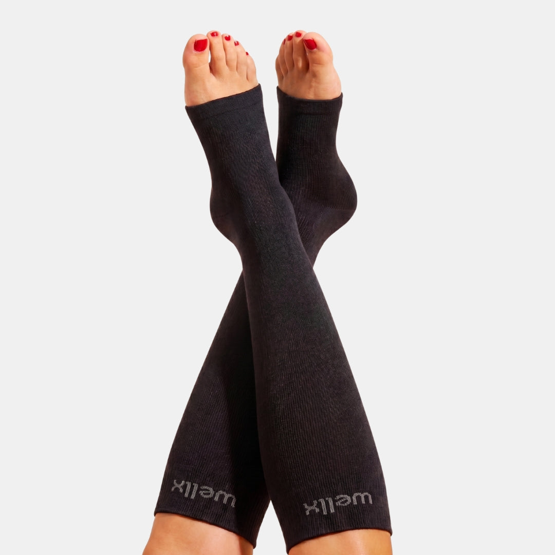 Black Toeless Compression Socks For Women Black ribbed compression calf sleeves worn on both legs, shown against a light gray background with the feet crossed. The brand name 'wellx' is visible in gray near the lower cuffs.