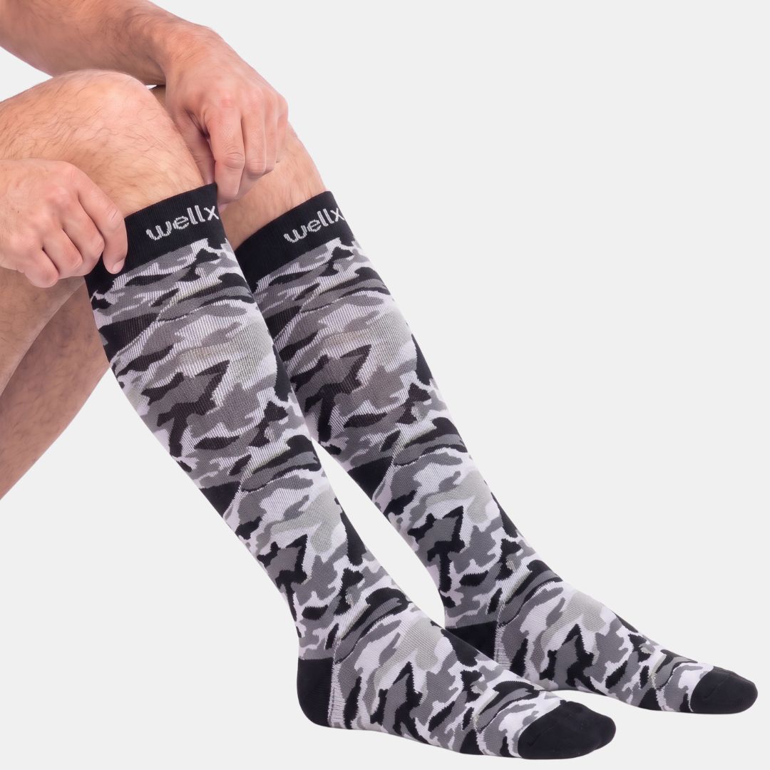 A pair of Stars Black Camouflage Compression Socks For Men featuring a colorful star camouflage pattern with teal cuffs, worn on legs against a light gray background. The Wellix brand name is visible on the cuff.