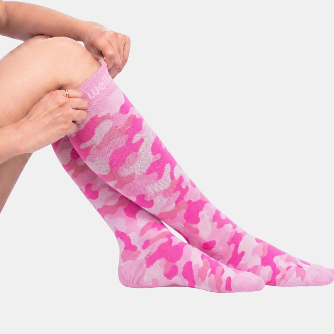 Camouflage Pink Compression Socks For Women, Pink camouflage knee-high socks worn on a model's legs, with the model's hands adjusting the top cuff. The brand name 'jeans' is partially visible in white text on the pink ribbed top band.