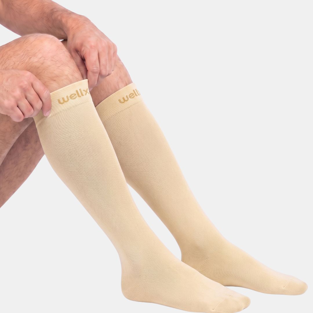 Candlelight Yellow Compression Socks For Men.  Beige knee-high compression socks worn on a person's legs, shown against a plain light gray background. The brand name 'wellx' is visible in gold on the top cuff of each sock.
