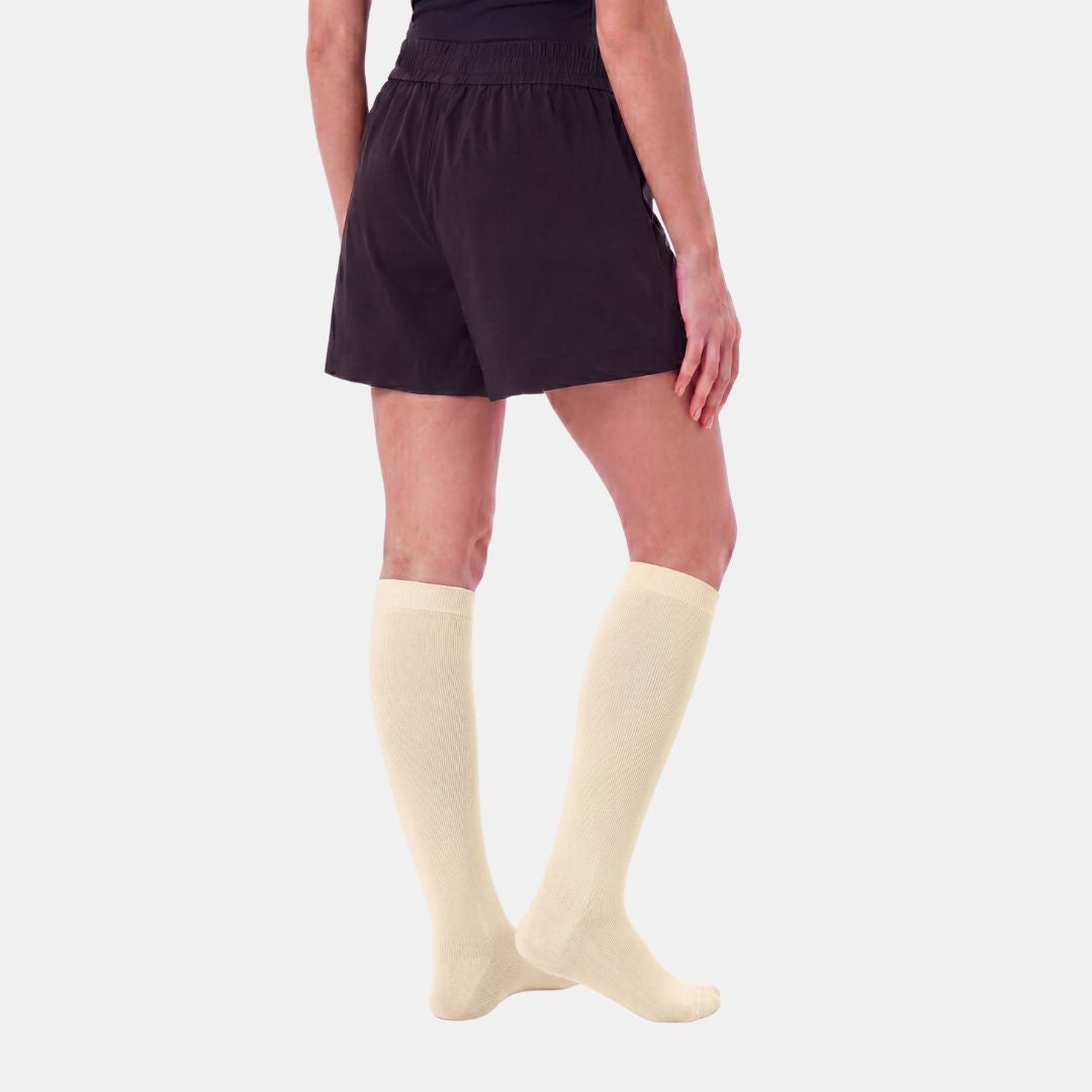 Candlelight Yellow Compression Socks For Women, A model from the back wearing black shorts and cream-colored knee-high socks against a light gray background.