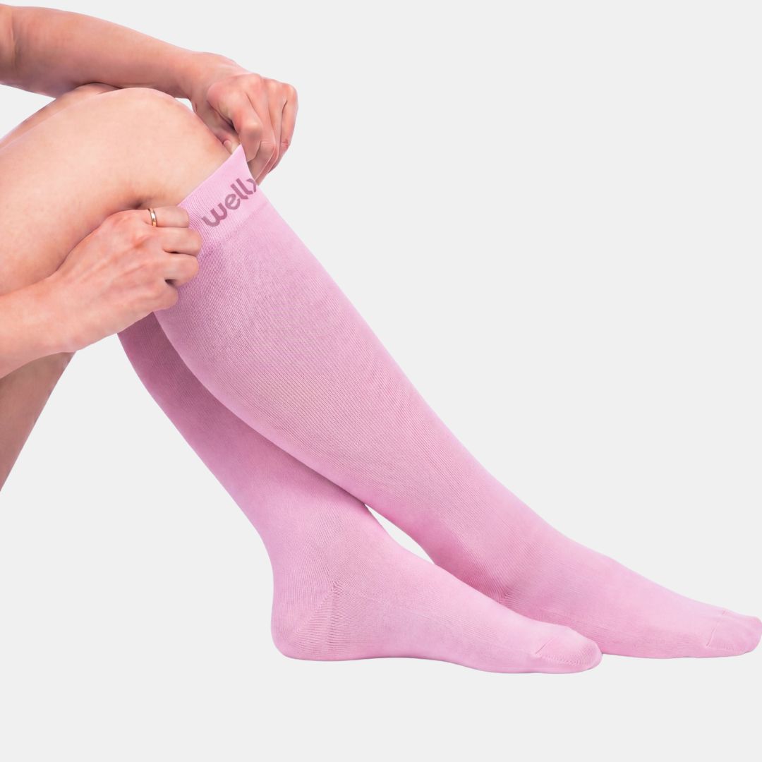 Candy Heart Pink Compression Socks For Women, A person wearing light pink over-the-knee socks, with their hands adjusting the top cuff. The brand name 'wellb' is visible in dark pink text on the top band.
