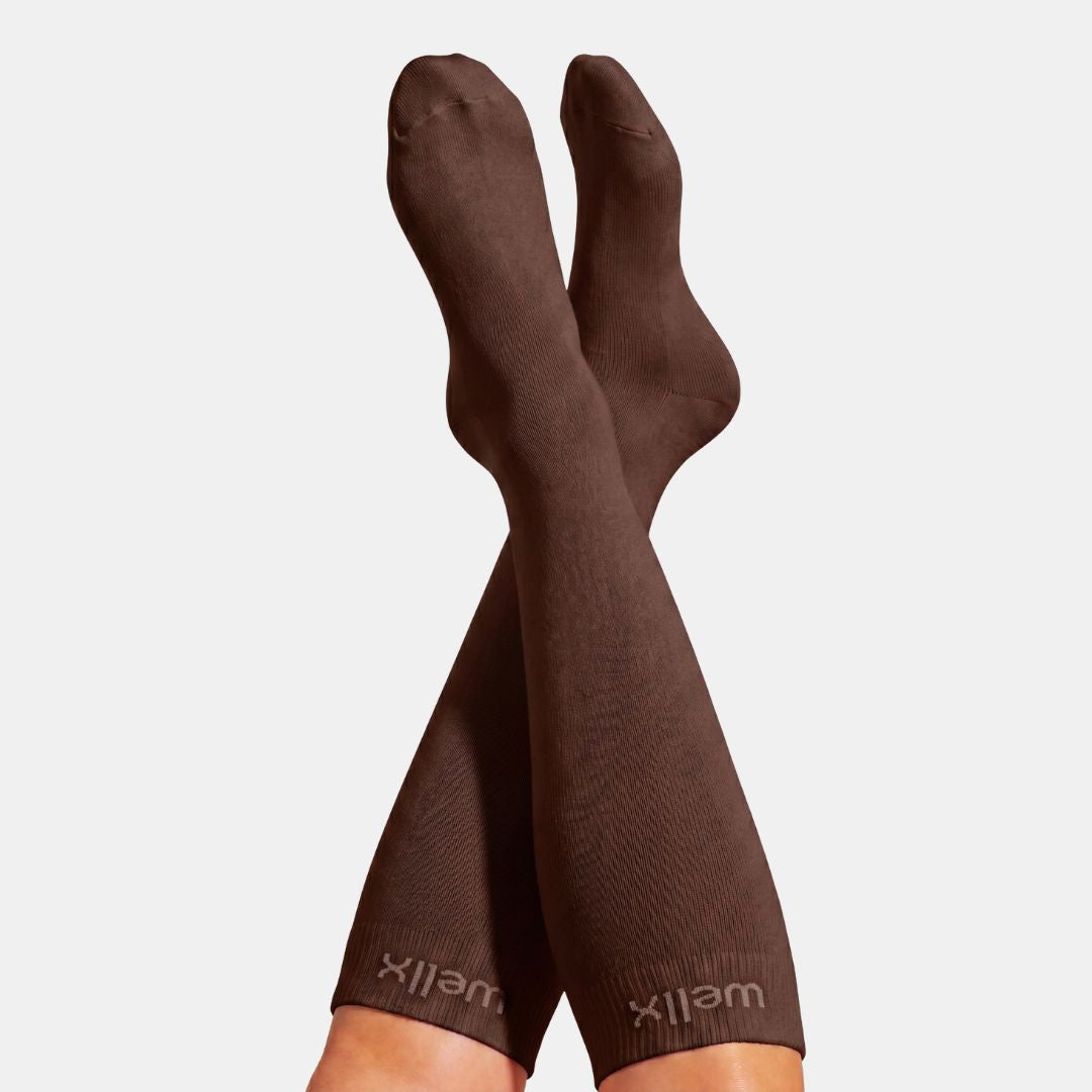 Espresso Compression Socks For Men, A pair of dark brown ribbed knee-high socks worn on crossed legs, with the brand name 'wellx' printed near the cuffs, against a light gray background.