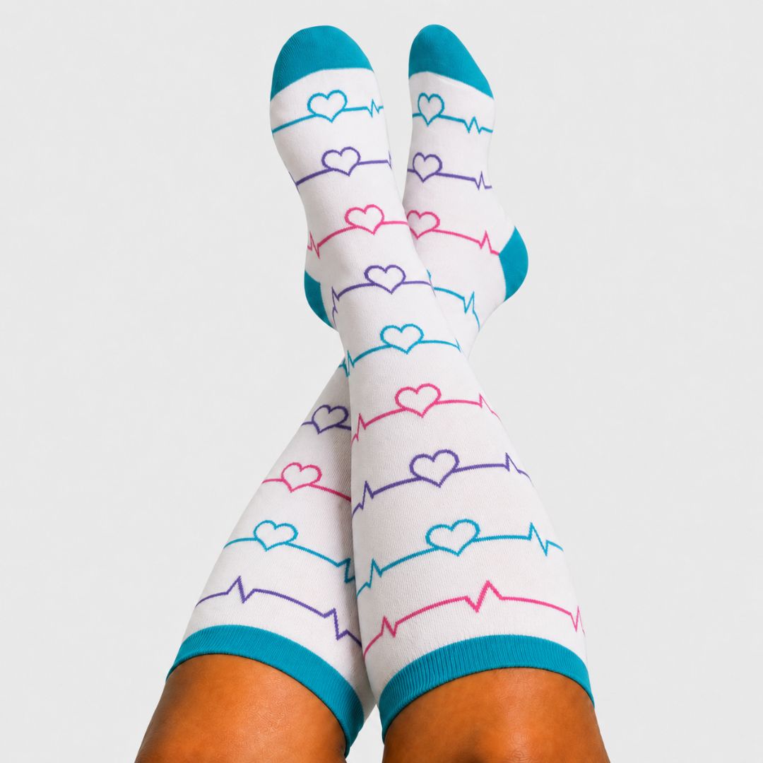 Heartbeat White Wide Calf Compression Socks For Men. White knee-high socks with a pattern of blue, pink, and purple heartbeat lines and hearts, featuring teal toes and cuffs, shown on a person's legs against a light gray background.
