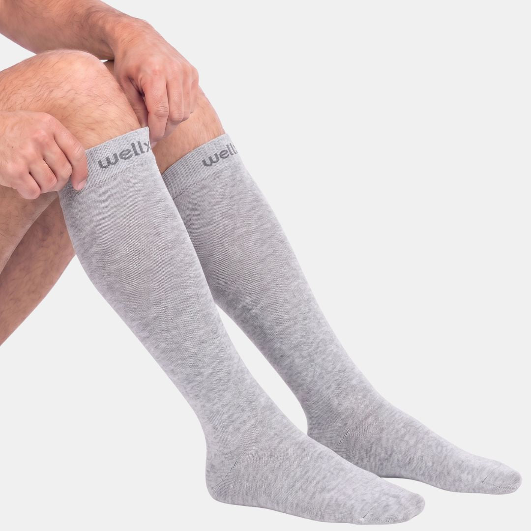 Silver Grey Compression Socks For Men,  A person wearing light gray heathered knee-high socks, with both legs visible against a plain light gray background. The socks have a ribbed texture and the brand name 'wellbo' is printed on the top cuff.
