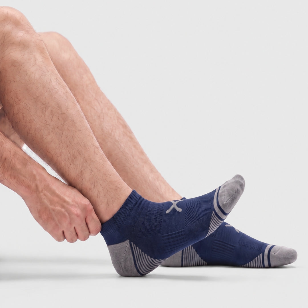 Navy Power Stride Ankle Socks Form Men. A close-up of a person's legs and feet wearing low-cut athletic socks in navy blue and gray, with a small white logo on the side, against a light gray studio background.