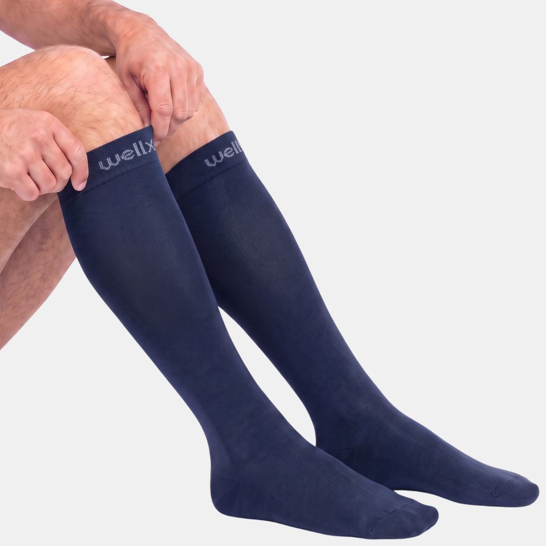 Navy Blue Compression Socks For Men, A close-up of a person's legs wearing dark navy knee-high compression socks with the brand name 'wellx' visible on the top cuff, set against a plain light gray background.