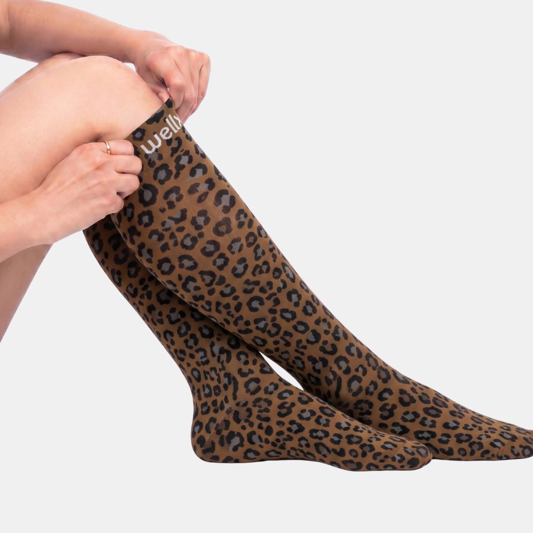 Panther Compression Socks For Women, A person wearing brown leopard-print knee-high socks, with one leg bent and the other sock being adjusted. The brand name 'wellit' is visible in white text on the cuff.