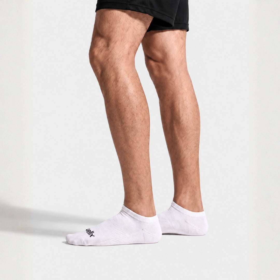 White Street Ankle Socks For Men. A close-up of a person's legs from the mid-thigh down, wearing black athletic shorts and white low-cut ankle socks with a small black logo on the toe, set against a clean white studio background.