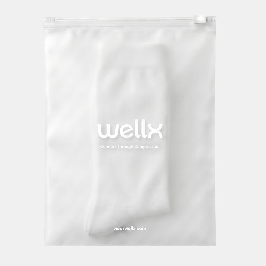 White Wide Calf Compression Socks For Men. A white compression garment, likely a stocking or sleeve, neatly folded and sealed inside a clear, zip-top plastic storage bag. The bag features the 'wellx' brand logo printed in white in the center, with 'wearwellx.