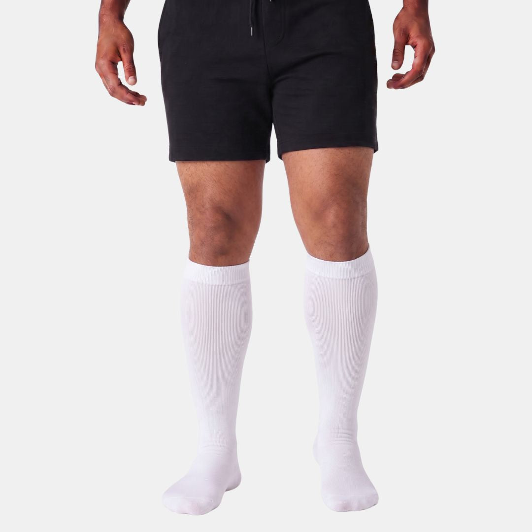 White Wide Calf Compression Socks For Men A model from the waist down wearing black athletic shorts and white knee-high compression socks against a light gray background.